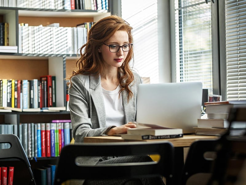 A woman working on a laptop at a desk in front of a full bookshelf
