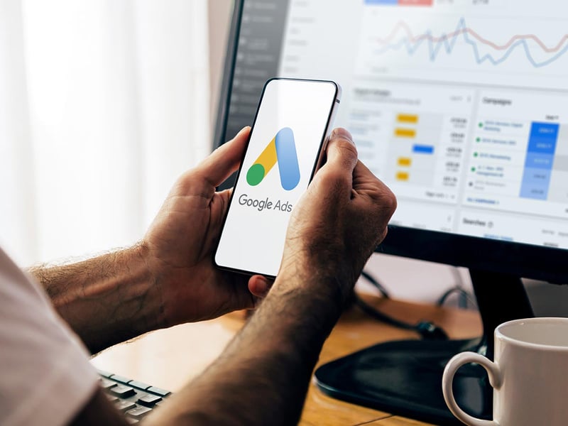 Closeup of hands using a mobile phone with Google Ads on the screen, and in the background, a computer showing ad statistics