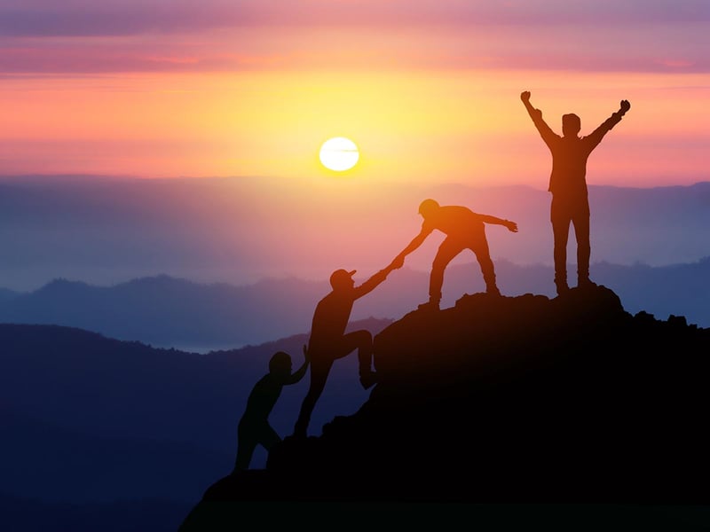 Silhouettes of four people helping each other climb a mountain at sunset