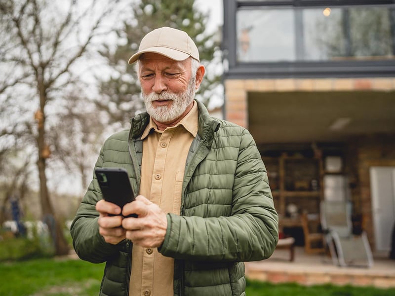 A senior man stands in front of a tiny house in the day, using a smartphone